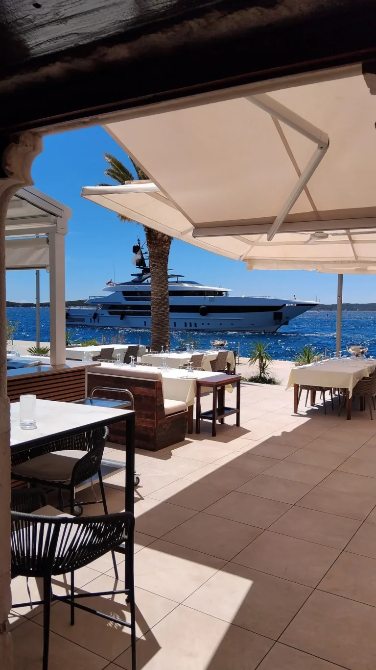 Shaded outdoor terrace of Gariful restaurant overlooking the bright blue sea, with a large luxury yacht anchored just offshore on a sunny day