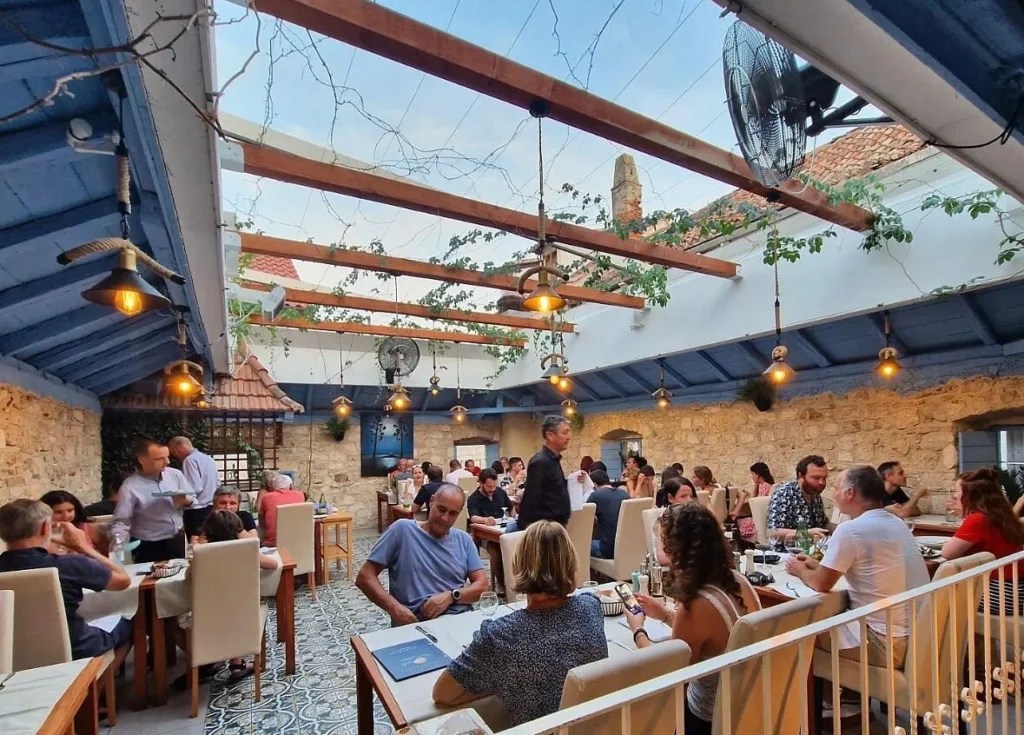 Bustling open-air dining area at Restaurant Grande Luna, featuring stone walls, wooden beams with hanging lights, patterned floor tiles, and guests enjoying dinner in a cozy Mediterranean atmosphere