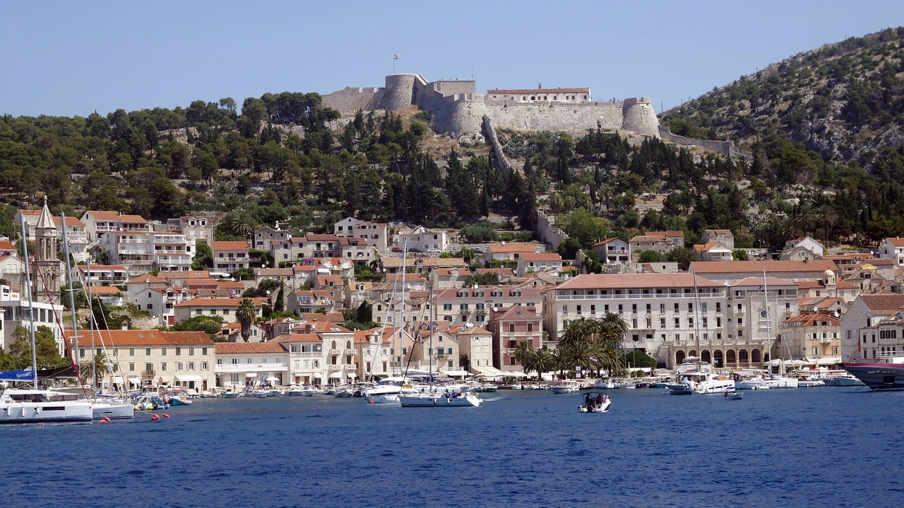 Waterfront view of Hvar town with stone buildings, red-tiled roofs, a lively marina full of boats, and the historic Fortica fortress rising above the town on a forested hill