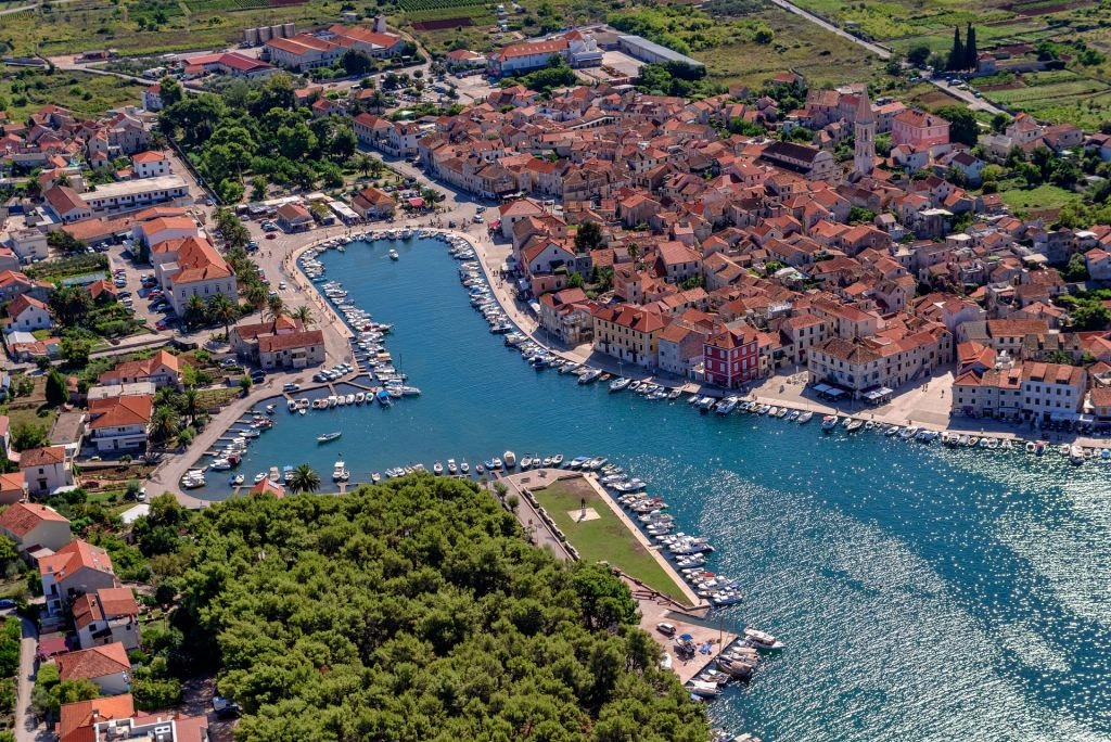 Aerial view of Stari Grad on Hvar, featuring a sheltered harbor lined with boats, densely packed stone houses with red-tiled roofs, and surrounding greenery under a bright sky