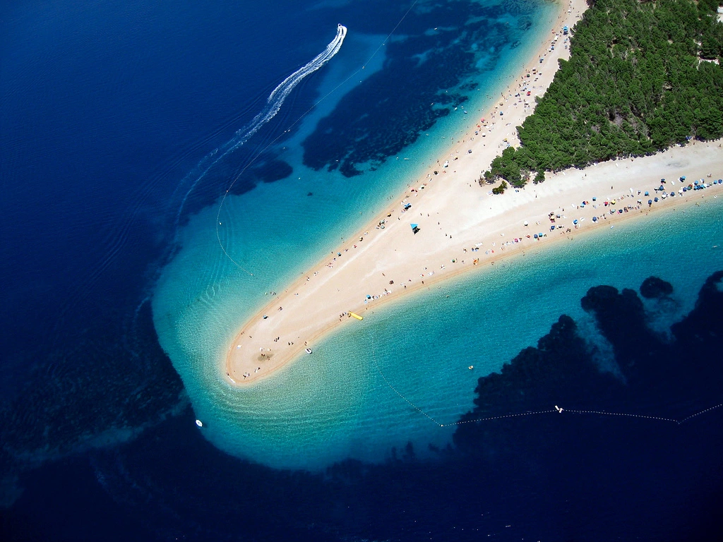 Aerial view of Zlatni Rat beach on the island of Brač, showing its iconic narrow pebble cape stretching into the turquoise and deep blue sea, with visitors scattered along the shoreline