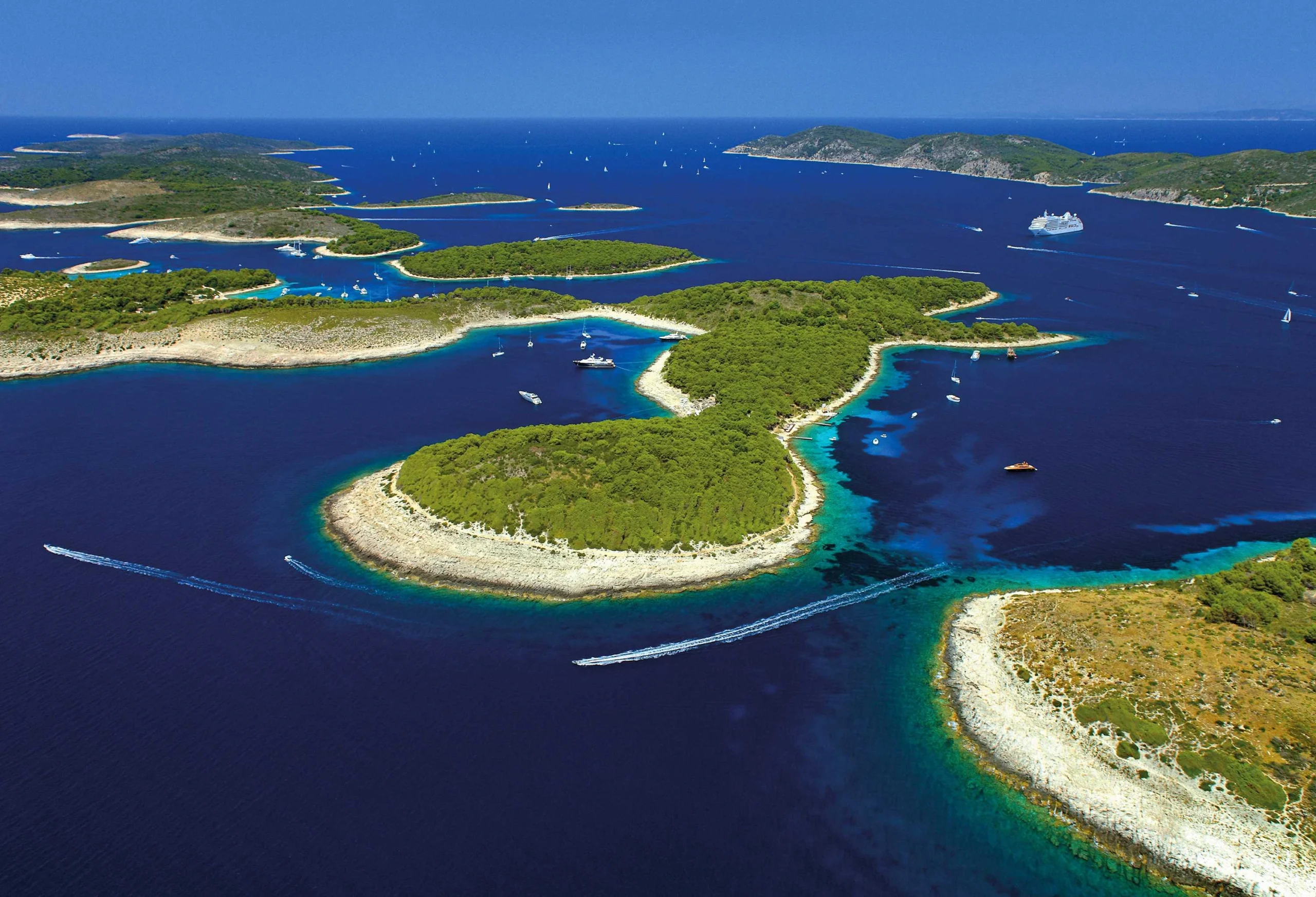 Aerial view of the Pakleni Islands near Hvar, showing a chain of lush green islets surrounded by deep blue sea, turquoise shallows, and boats scattered across the water