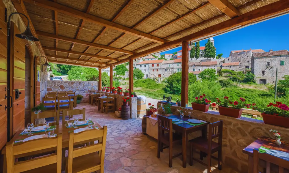 Tradycyjna kamienna wioska Velo Grablje na wyspie Hvar widziana z rustykalnej restauracji z tarasem, drewnianymi stołami i górskim tłem
