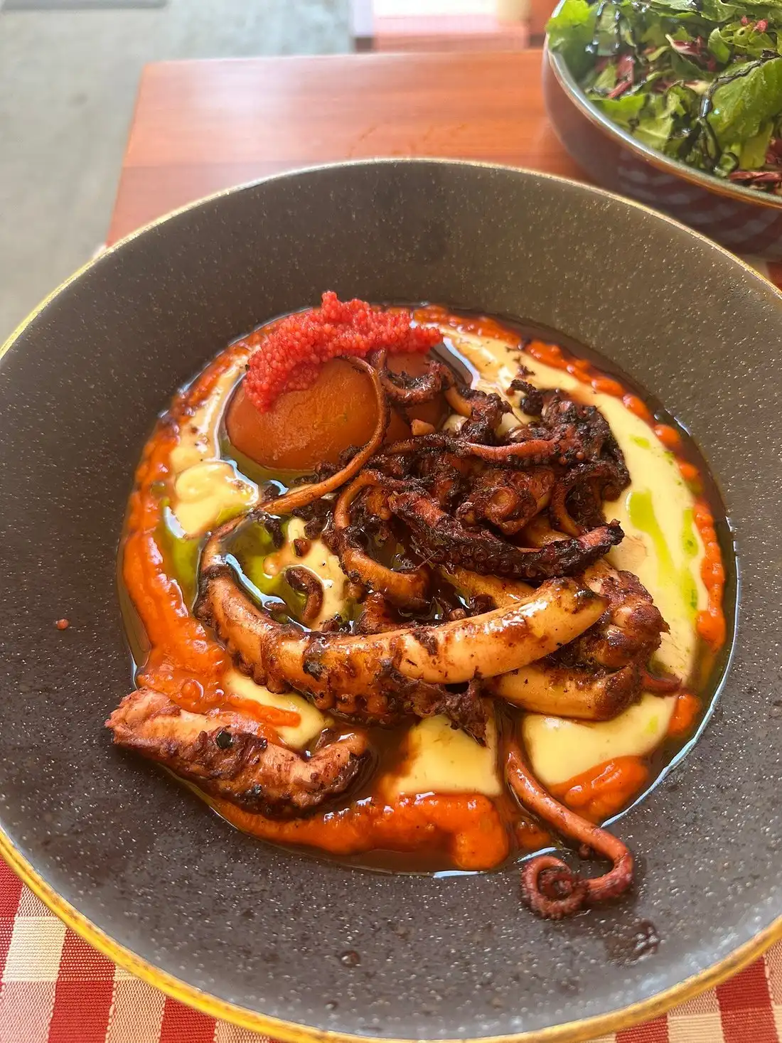Grilled octopus served over creamy mashed potatoes with red pepper sauce and roe, presented in a rustic black bowl at a restaurant