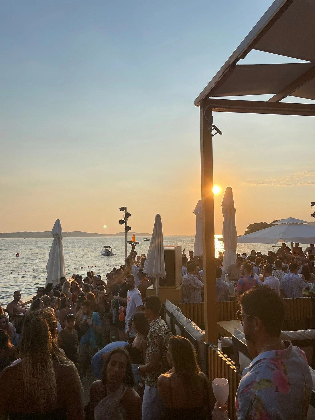 Crowded beach bar at sunset, with people dancing and socializing near the sea as the sun sets over the water