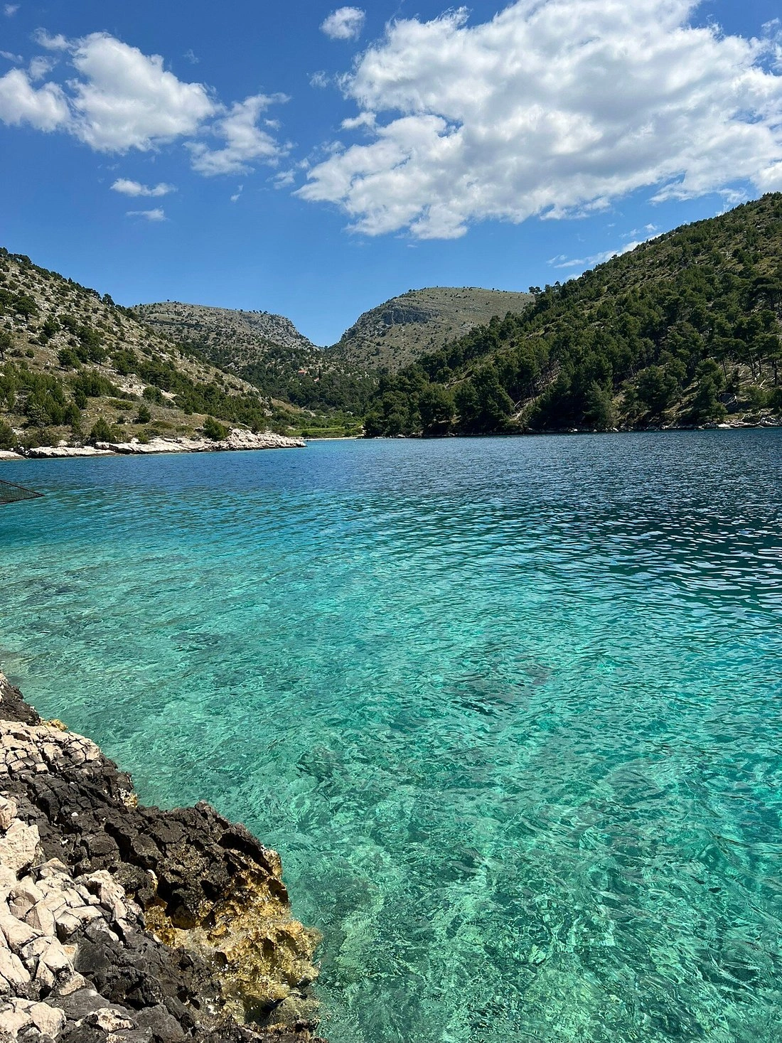 Baie turquoise aux eaux cristallines, entourée de rivages rocheux et de collines verdoyantes, sous un ciel d'un bleu éclatant parsemé de nuages.