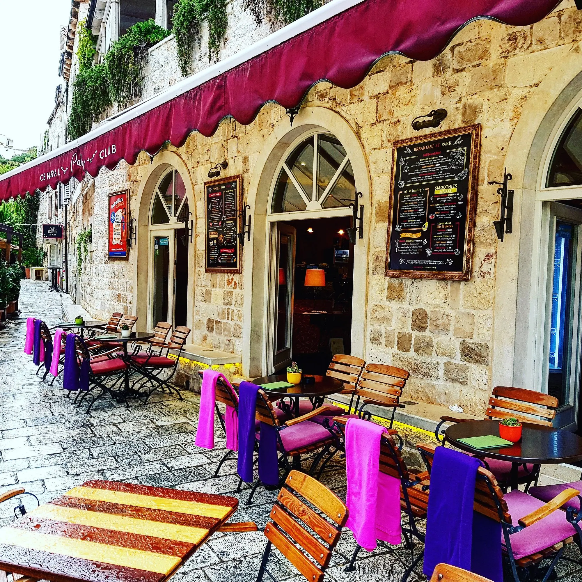 Colorful outdoor café seating with pink and purple blankets draped over wooden chairs along a stone-paved street in Hvar’s old town, outside the Central Park Club with its arched windows and red awning
