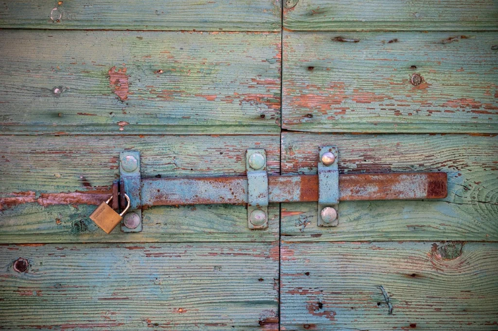 Close-up of an old, weathered wooden door painted in faded turquoise, with a rusty metal latch secured by a brass padlock