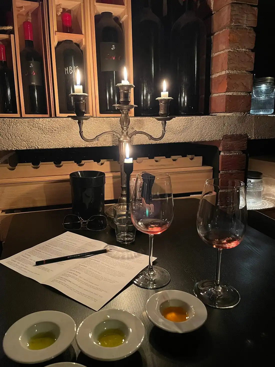 Candlelit wine tasting setup with two half-filled wine glasses, tasting notes, small dishes of olive oil, and large wine bottles displayed in a rustic cellar setting