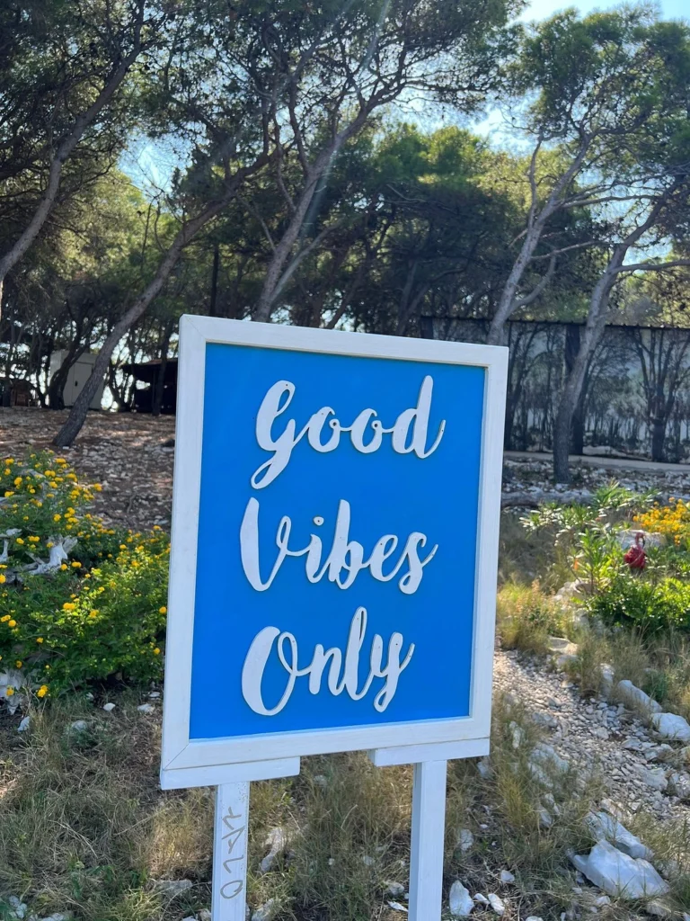 Une pancarte bleue avec des lettres blanches cursives sur lesquelles on peut lire « Good Vibes Only » est placée à l'extérieur, parmi les arbres, les rochers et les fleurs jaunes, par une journée ensoleillée.