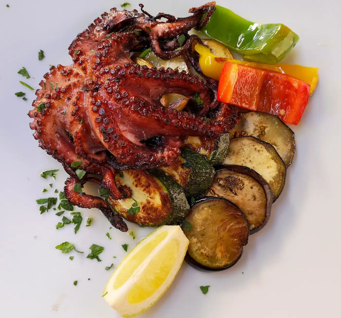 Grilled octopus served with roasted zucchini, eggplant, and colorful bell peppers, garnished with fresh parsley and a lemon wedge on a white plate