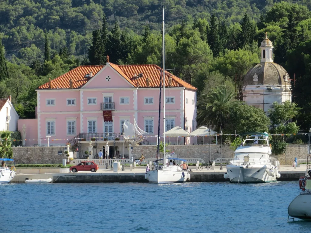 Waterfront view of Heritage Villa Apolon in Stari Grad, a pink historic building with a red-tiled roof, surrounded by greenery and boats docked along the harbor