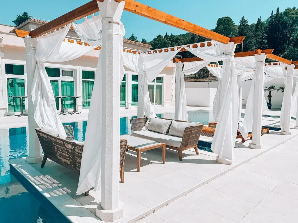 Elegant poolside cabanas with white curtains and wicker lounge seating at Hotel Antica, set beside a turquoise swimming pool with surrounding trees in the background