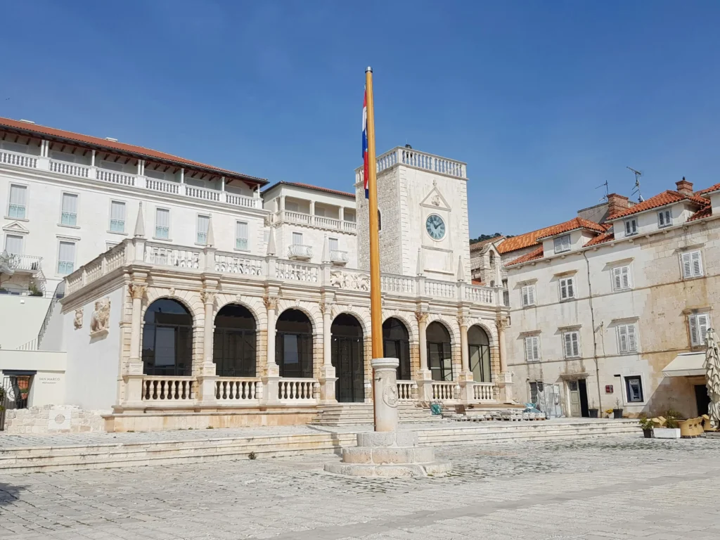 Historic stone facade of the Palace Elisabeth Heritage Hotel in Hvar, featuring arched loggia columns, a central clock tower, and a Croatian flagpole on the main town square under a clear blue sky.