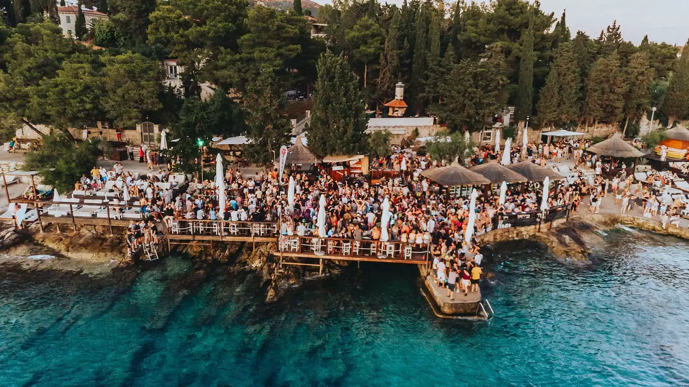 A lively seaside crowd gathered on wooden decks above turquoise water at a popular Hvar beach bar surrounded by pine trees