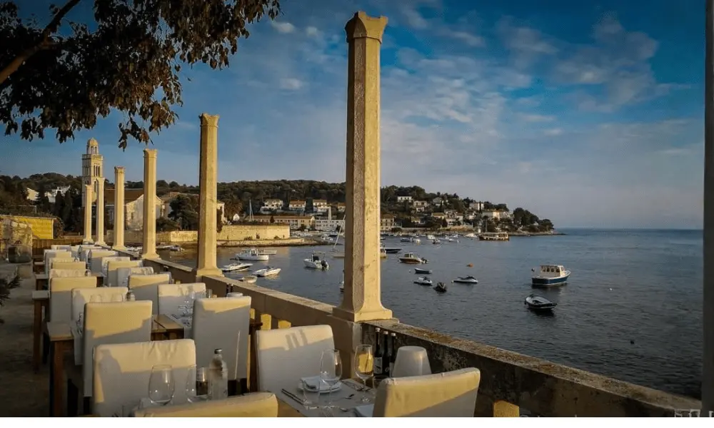 Taras restauracji z widokiem na morze w Hvarze, eleganckie stoliki i widok na port, łodzie i zabytkowe kamienne budynki o zachodzie słońca.
