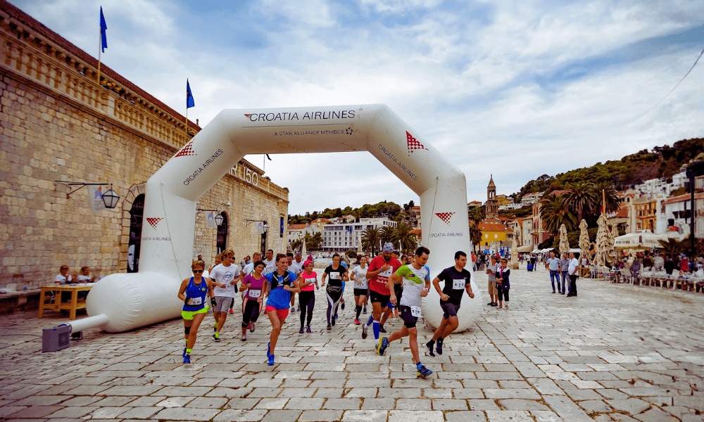 Biegacze biorący udział w biegu na historycznym placu miasta Hvar, przechodzący pod łukiem wydarzenia Croatia Airlines.