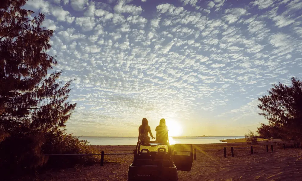 Dwóch przyjaciół siedzi w samochodzie terenowym i ogląda piękny zachód słońca nad plażą, otoczeni drzewami i otwartym niebem.