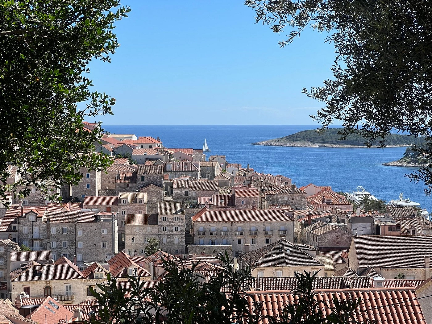View of Hvar’s old town with stone houses and terracotta rooftops framed by trees, overlooking the blue Adriatic Sea and nearby Pakleni Islands on a clear sunny day