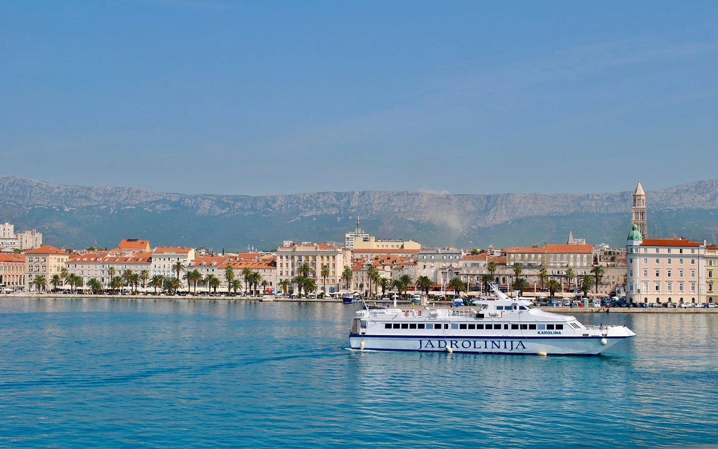 Jadrolinija ferry sailing in front of Split’s waterfront, with the Riva promenade lined with palm trees and the historic city skyline backed by the Marjan hills