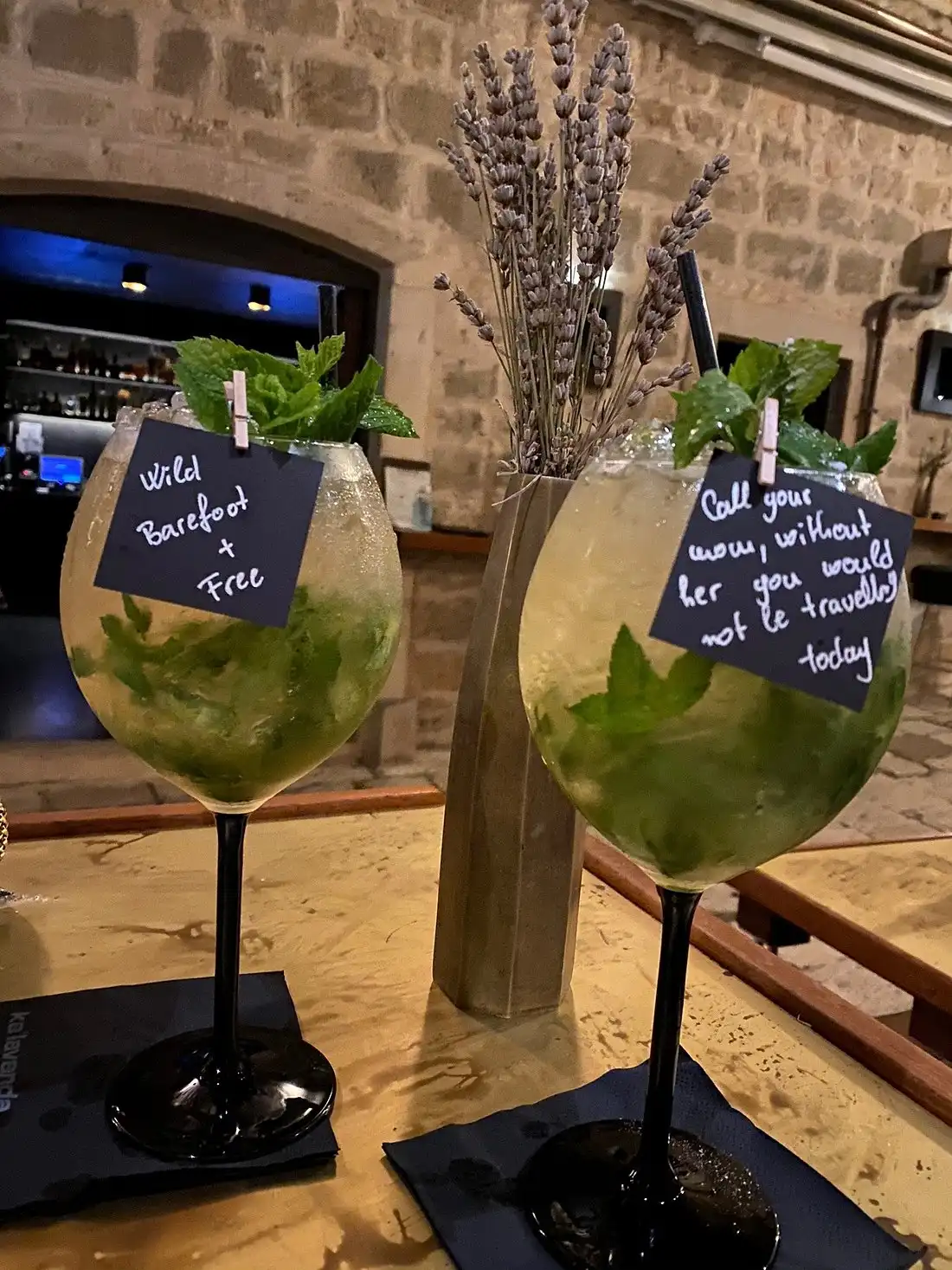 Two large cocktails garnished with fresh mint and small handwritten quote cards, placed on a bar table with dried lavender in the background inside a rustic stone-walled venue