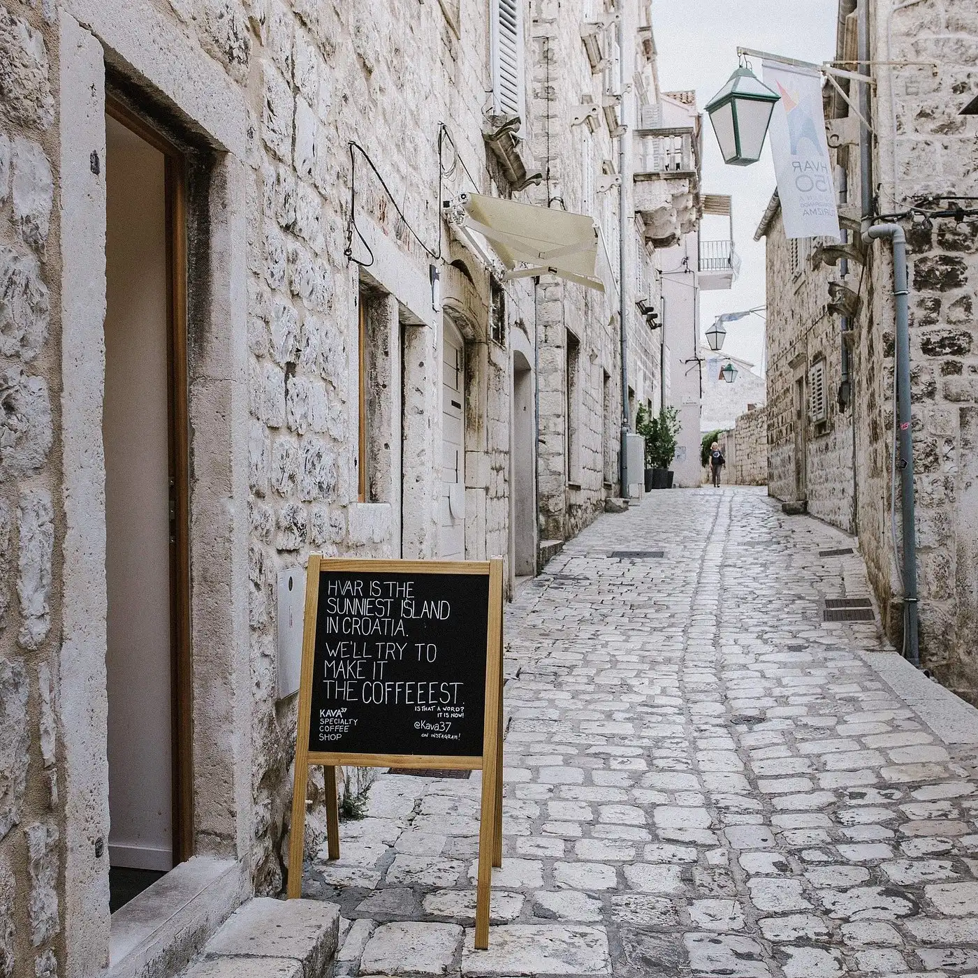 A narrow stone alley in Hvar’s old town with a chalkboard sign outside a café that reads ‘Hvar is the sunniest island in Croatia. We’ll try to make it the coffeeeest,’ surrounded by historic limestone buildings