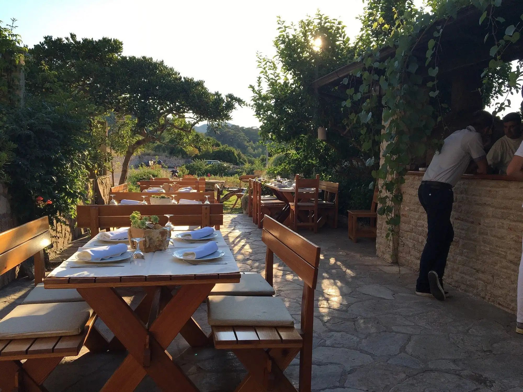 Outdoor terrace of a rustic Hvar restaurant at sunset, with wooden tables set for dinner, surrounded by lush greenery and stone walls as a few people chat at the bar