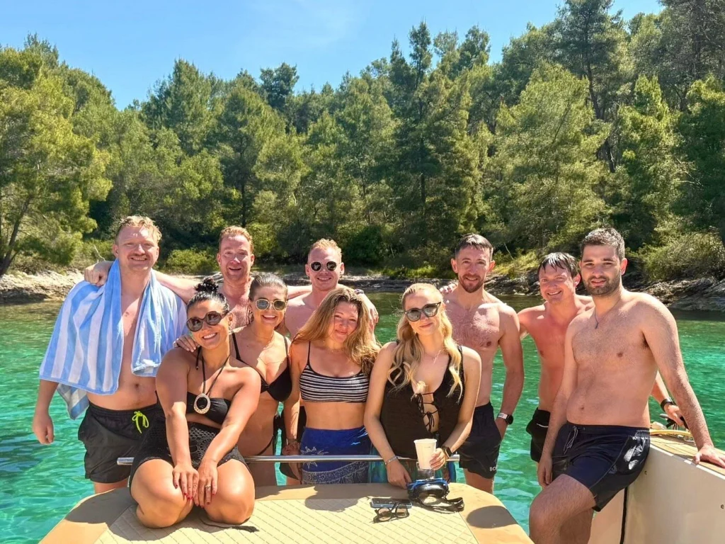 Group of friends posing and smiling on a boat in a turquoise cove surrounded by pine forest, enjoying a sunny summer day on the Adriatic Sea