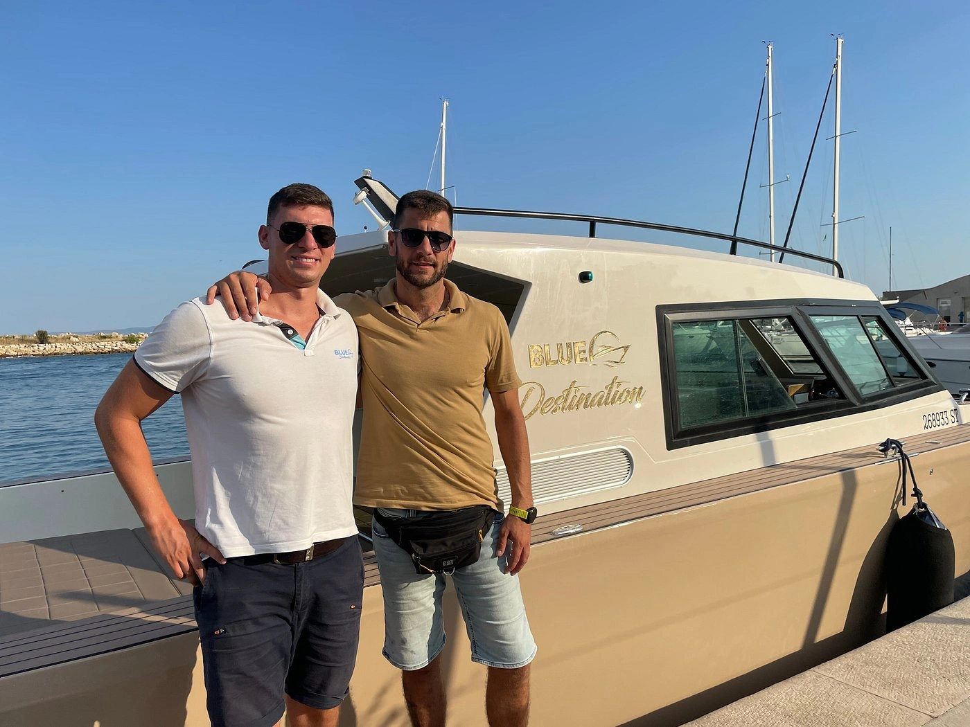 Two men standing together and smiling beside a boat at a marina on a sunny day.