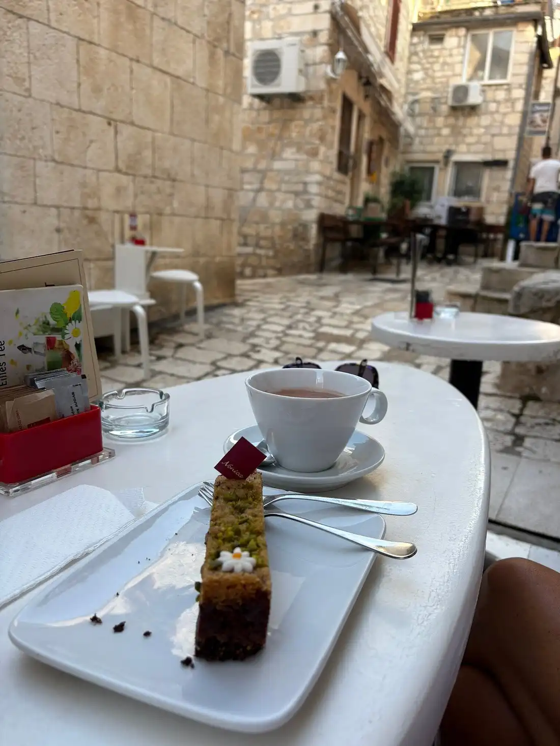 A slice of cake and a cup of coffee on a white café table set along a narrow stone alley in Hvar’s old town, surrounded by rustic limestone buildings and outdoor seating