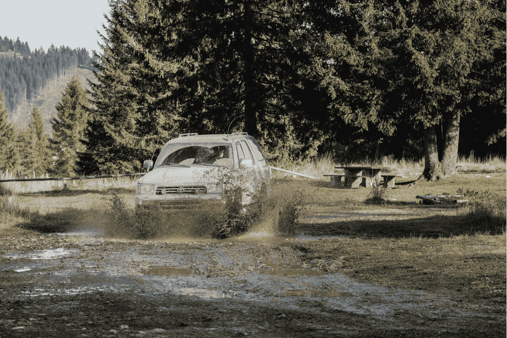 Off-road vehicle driving through a muddy puddle in a forested area, splashing water and mud