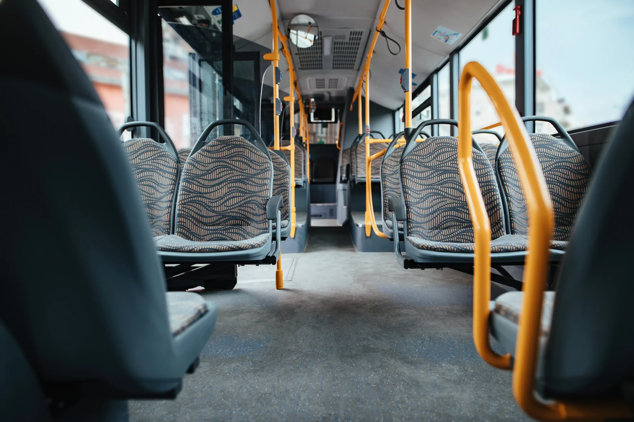 Empty interior of a public bus with patterned seats and yellow handrails, viewed from a low angle looking down the aisle