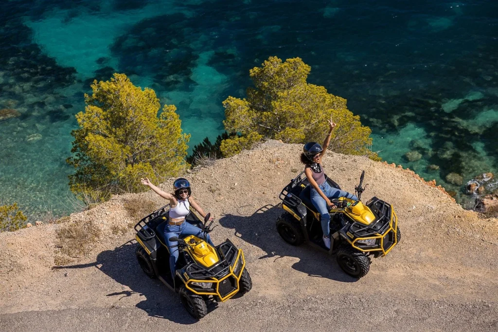 Deux femmes à bord de quads jaunes longent une route côtière escarpée, souriantes et posant les bras levés au-dessus de la mer Adriatique turquoise.