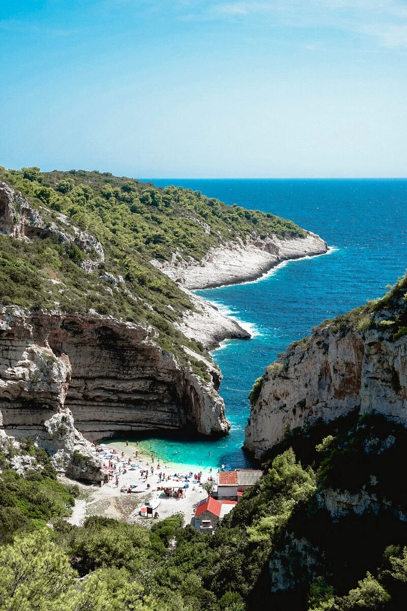 Scenic view of Stiniva Cove on the island of Vis, showing a narrow rocky inlet opening into turquoise water, surrounded by steep cliffs and green vegetation, with a small pebble beach filled with sunbathers below