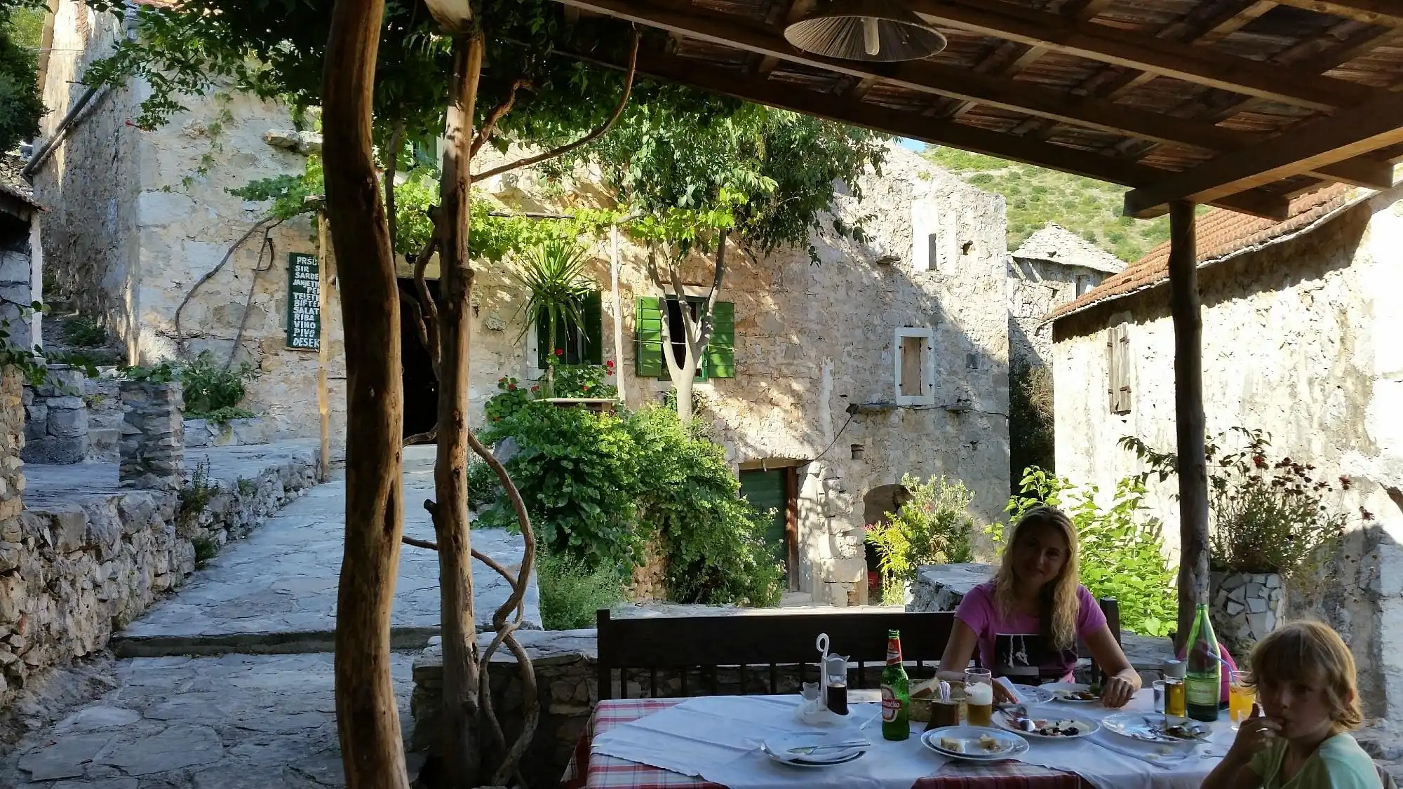 A rustic outdoor dining table in a traditional stone village on Hvar, with a woman and child enjoying a meal under a shaded terrace surrounded by lush greenery and historic stone houses