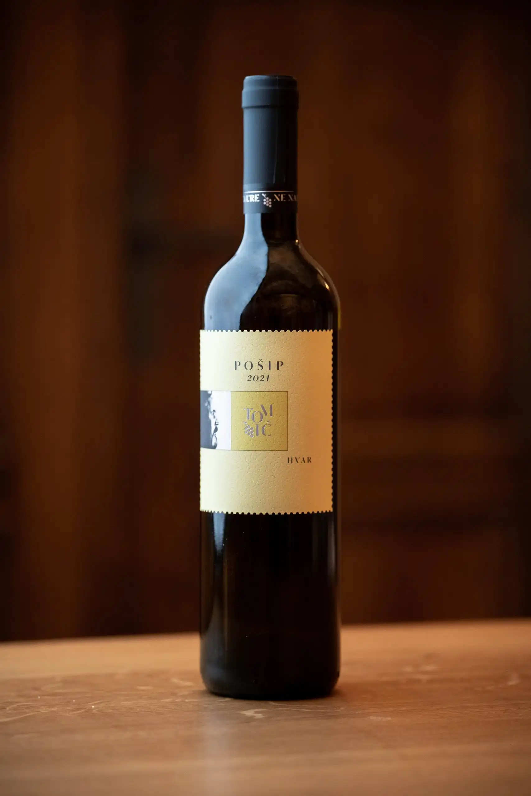 A bottle of Tomic Pošip 2021 white wine from Hvar displayed on a wooden table with a softly blurred background.