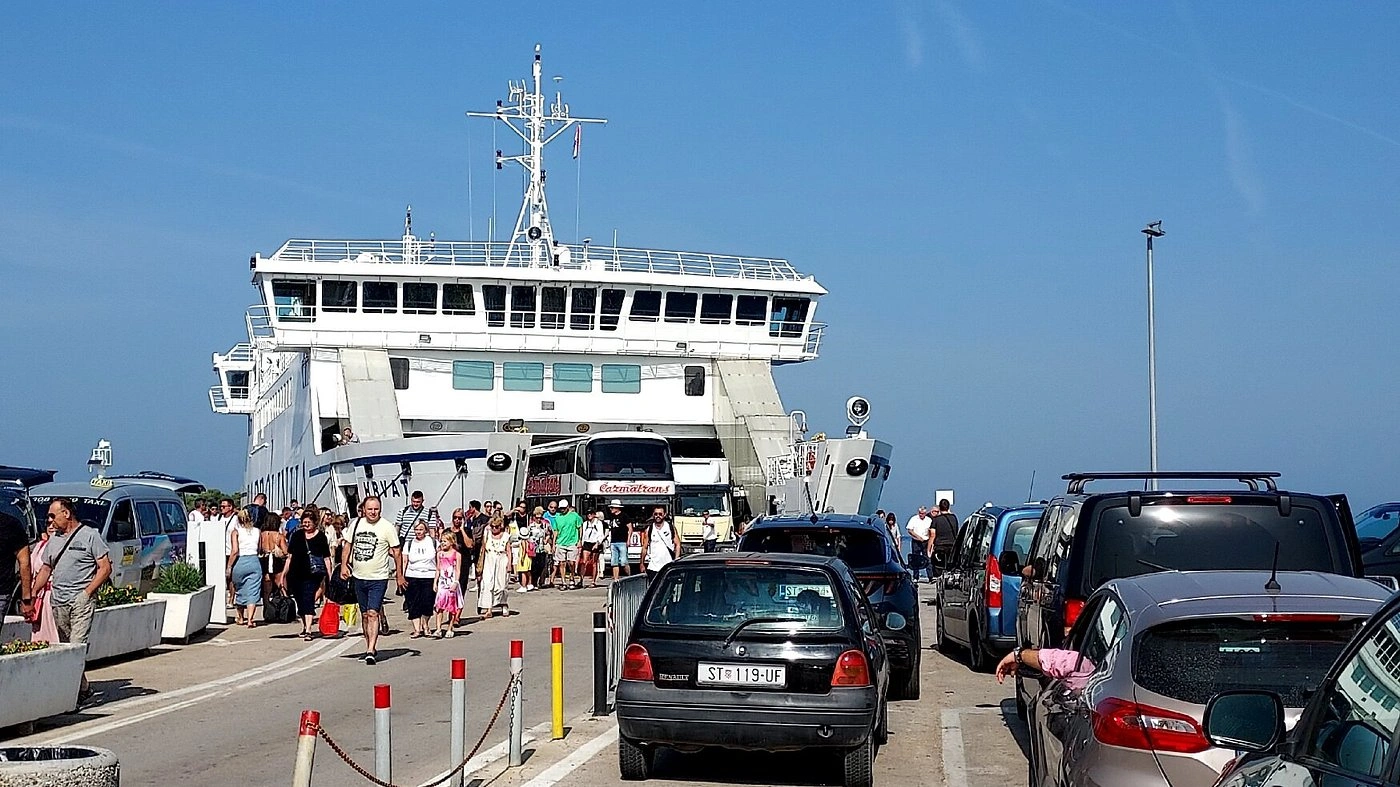 Une foule de passagers et une file de voitures embarquent ou débarquent d'un grand ferry dans un port animé par une journée ensoleillée.