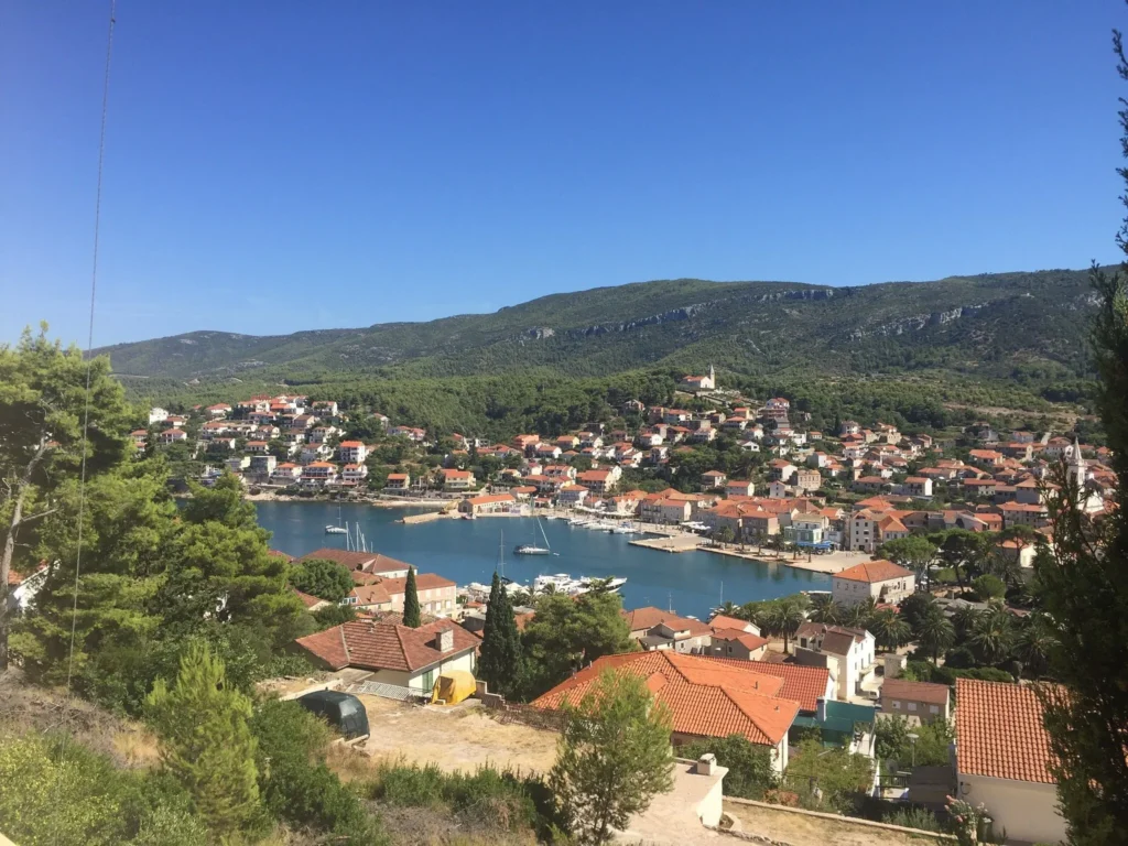 Panoramic view from the terrace of Villa Vista overlooking Jelsa, with red-roofed houses, a calm harbor dotted with boats, and forested hills under a clear blue sky.