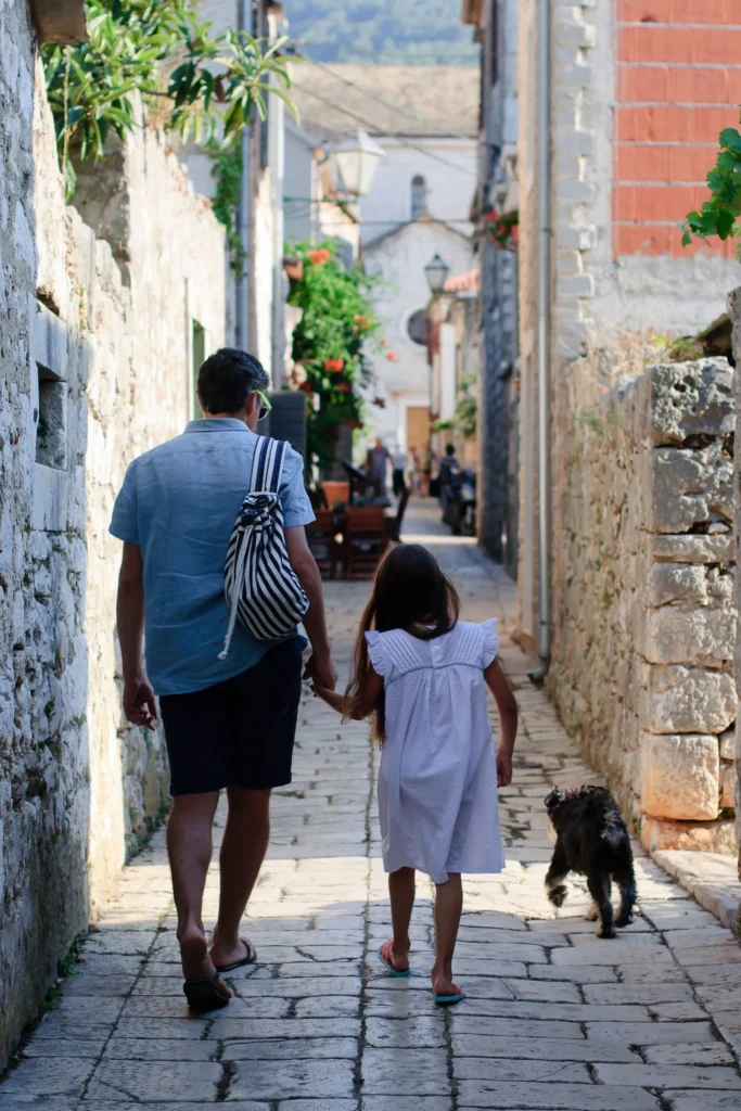 A man holding a young girl’s hand as they walk with a small dog through a narrow stone street in a historic Mediterranean town
